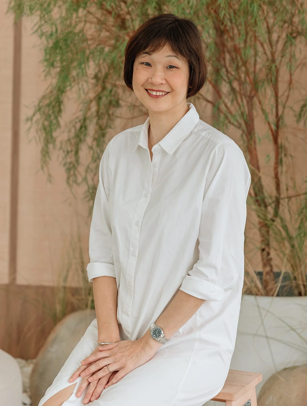 A smiling woman with short dark hair in a white shirt sits in front of a large leafy green plant on a beige wall.